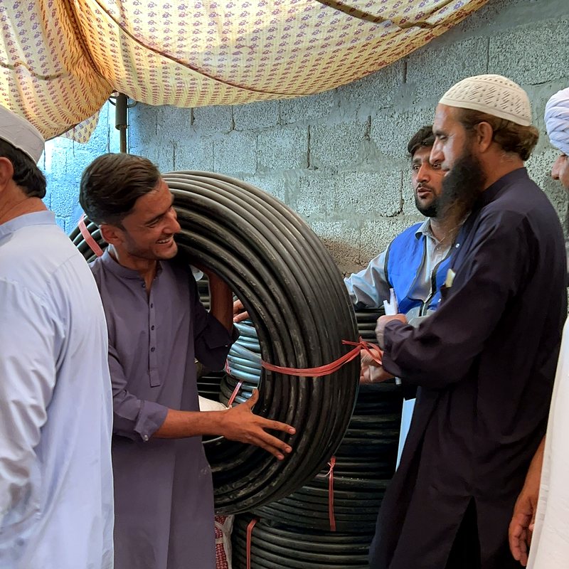 A smiling man shouldering a coil of water pipe while volunteers coordinate the delivery