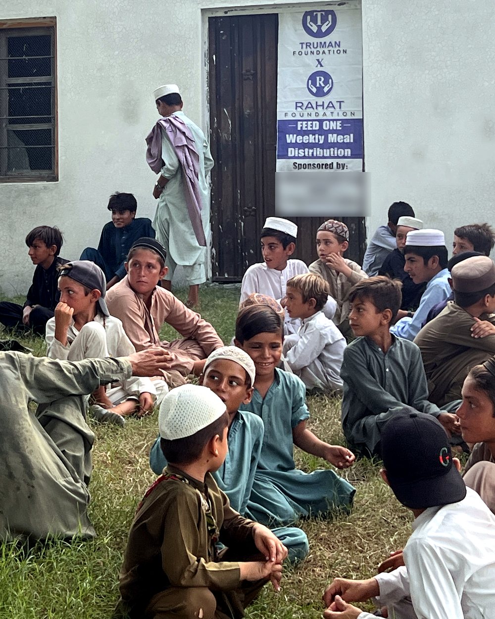 Village boys gathered on the grass before a Feed One meal, one boy smiling brightly beneath the Truman × Rahat banner